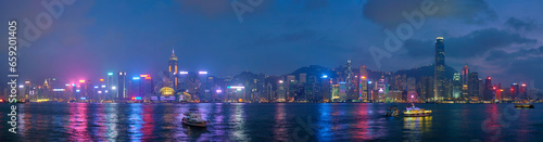 Photography Panorama of Hong Kong skyline cityscape downtown skyscrapers over Victoria Harbour in the evening illuminated with tourist boat ferries