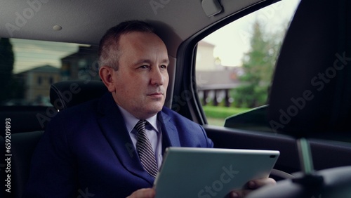 Lawyer man works in car. Serious businessman uses computer tablet to obtain business information in interior of business car. Politician works and looks at information on tablet in car. Man in suit