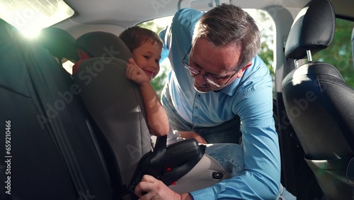 Dad puts her Kid boy in car seat safe car. Happy family. Father secures her kid son in car seat using child safety belt. Family road trip Dad cares about her son safety. Child sits in car child seat