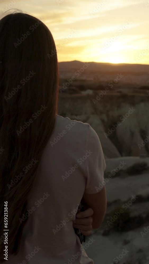 Vertical video. View from behind a young woman with long hair, the sun is almost setting behind the distant mountain, and she is enjoying the sunset. Vertical video. View from behind a young woman with long hair, the sun is almost setting behind the distant mountain, and she is enjoying the sunset.