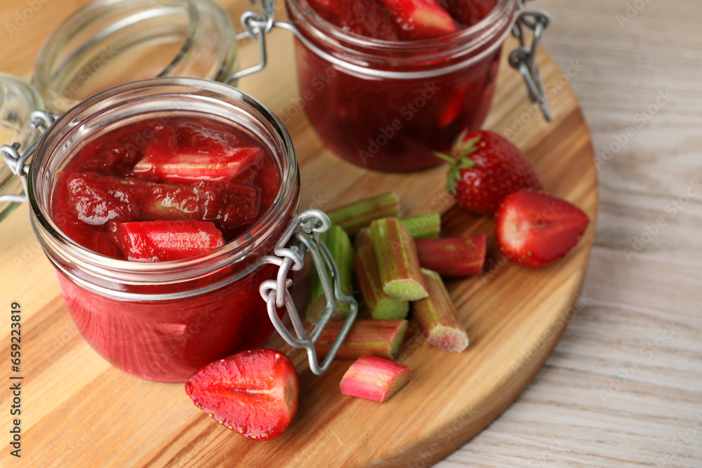 Jars of tasty rhubarb jam, cut stems and strawberries on white wooden table Jars of tasty rhubarb jam, cut stems and strawberries on white wooden table