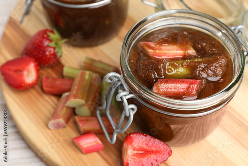 Jars of tasty rhubarb jam, cut stems and strawberries white wooden table, closeup