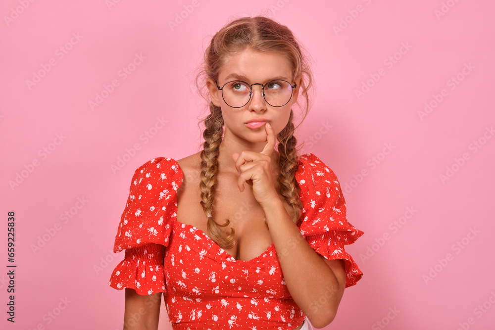 Young embarrassed pretty Caucasian woman teenager puzzledly looks to side and touches lips deciding which university to go to after graduating from school stands in pink studio. Student, pupil Young embarrassed pretty Caucasian woman teenager puzzledly looks to side and touches lips deciding which university to go to after graduating from school stands in pink studio. Student, pupil