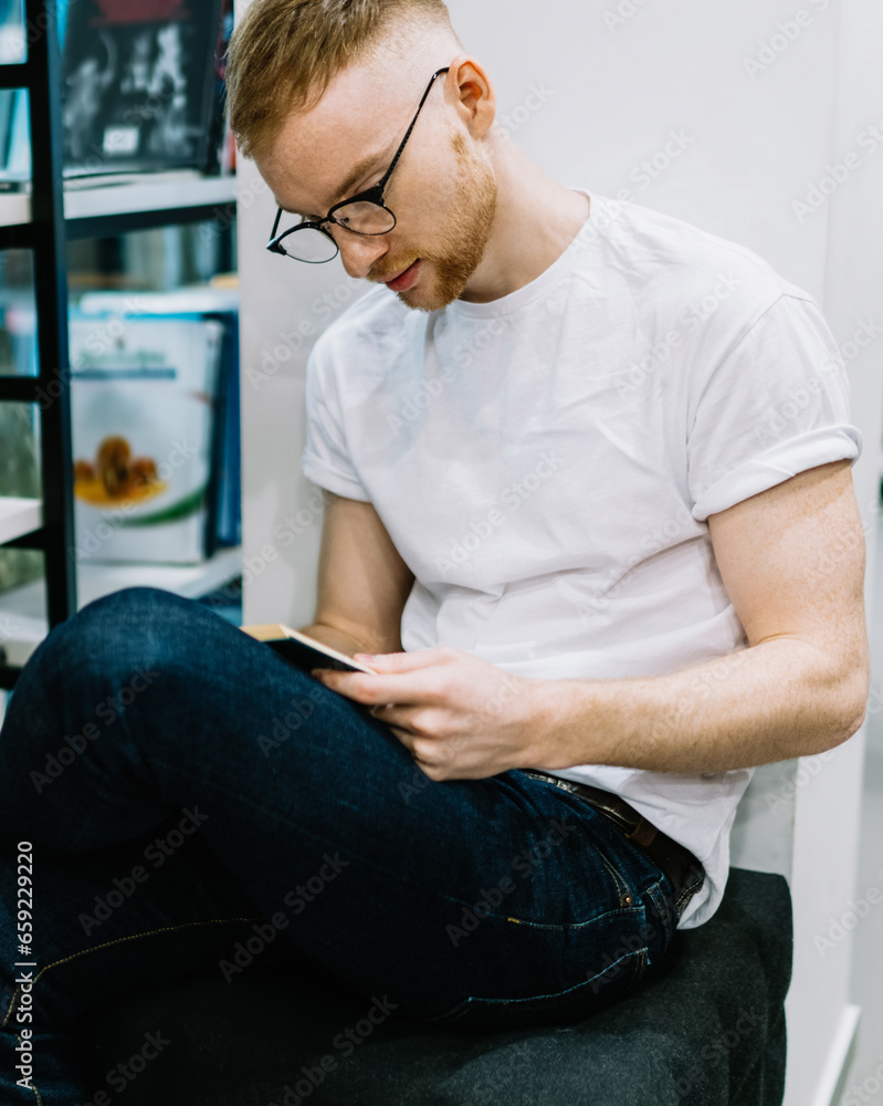 Thoughtful guy sitting cross legged and reading book in library Thoughtful guy sitting cross legged and reading book in library