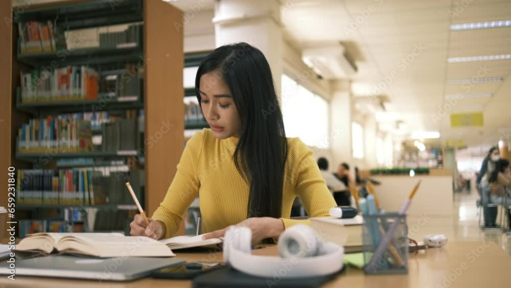 Asian student girl sitting at table looking book while do homework with laptop making video call abroad using internet friend connection, happy mood smiling broadly in library Asian student girl sitting at table looking book while do homework with laptop making video call abroad using internet friend connection, happy mood smiling broadly in library