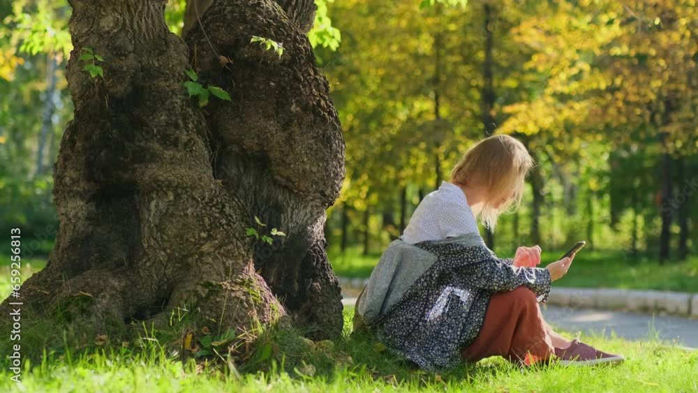 A pretty caucasian woman using her smartphone while sitting near big tree in an autumn park on a sunny day. A happy blonde is enjoying weekend outdoors. A pretty caucasian woman using her smartphone while sitting near big tree in an autumn park on a sunny day. A happy blonde is enjoying weekend outdoors.