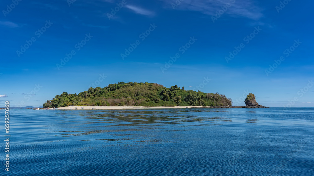 The island in the ocean is covered with green vegetation. Nearby is a picturesque rock. Tiny silhouettes of people, moored boats on the sandy beach. Blue shiny water, azure sky. Copy space. Madagascar
