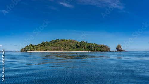 Fototapeta Naklejka Na Ścianę i Meble -  The island in the ocean is covered with green vegetation. Nearby is a picturesque rock. Tiny silhouettes of people, moored boats on the sandy beach. Blue shiny water, azure sky. Copy space. Madagascar