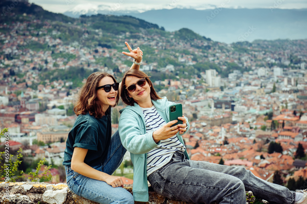 Two cheerful traveler girls in dark glasses are having fun and taking pictures against the background of Sarajevo, a beautiful European city in the mountains