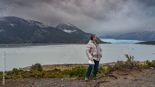 A man in a down jacket is standing on the shore of the lake, looking into the distance, to the side, smiling. In the distance, between the mountain slopes, the blue glacier Perito Moreno is visible. 