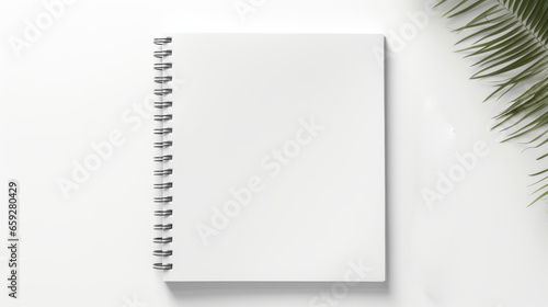 A clean and simple image of a blank spiral notebook on a white background with a green palm frond. The notebook is white with a silver spiral binding on the left side. The palm frond adds a touch of