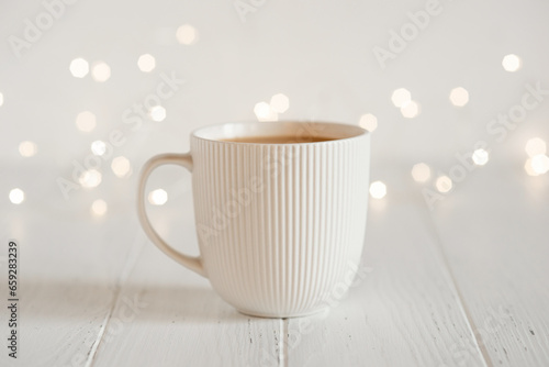 A white cup with hot coffee or tea on a white wooden table with Christmas lights.