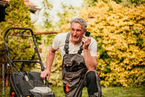 A mature man dressed in formal attire dealing with a malfunctioned lawnmower, resorting to a phone call to solve the issue
