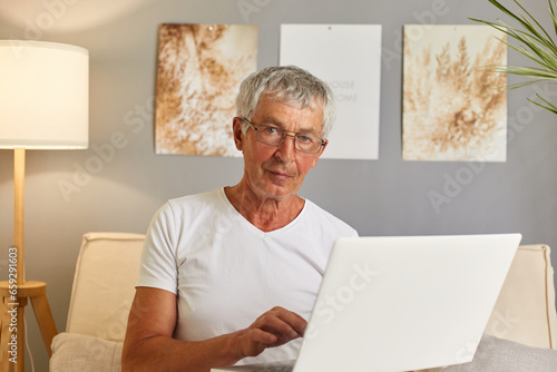 Positive grey-haired senior man in casual white T-shirt and glasses sitting on couch at home in living room interior using laptop answering e-mails typing massage browsing internet.