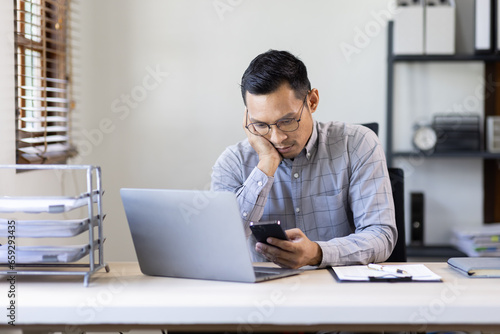 Portrait of tired young business Asian man work on phone with documents tax laptop computer in office. Sad, unhappy, Worried, Depression, or employee life stress concept