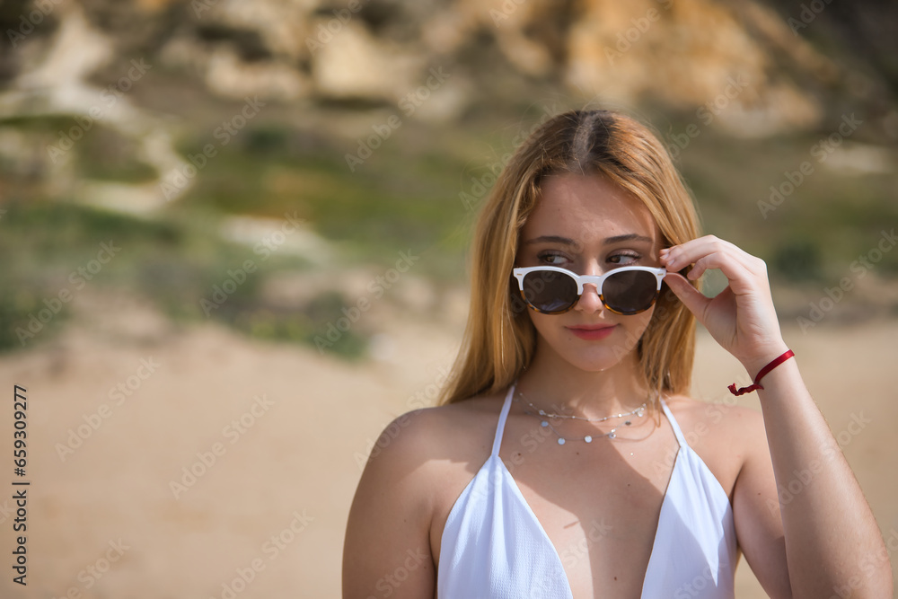 Young, pretty, blonde woman in a white dress, looking flirtatiously over sunglasses, on the beach. Concept sunglasses, beach, flirting. Young, pretty, blonde woman in a white dress, looking flirtatiously over sunglasses, on the beach. Concept sunglasses, beach, flirting.