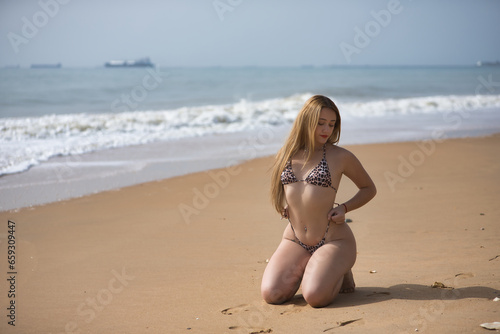 Young woman, blonde and beautiful, with a leopard print bikini, kneeling on the shore of the beach, in sensual and provocative attitude. Concept beach, bikini, sensuality, provocation. © Manuel