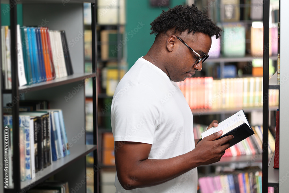 Confident young smart african student reading book in library wearing glasses. African American man in white t-shirt studying in library - obrazy, fototapety, plakaty 