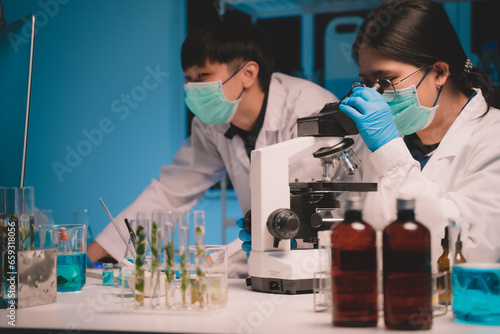 Two scientists looking through a microscope and recording an ecological skin care experiment in a night lab development concept. Research with plants and scientific extraction in glassware. 