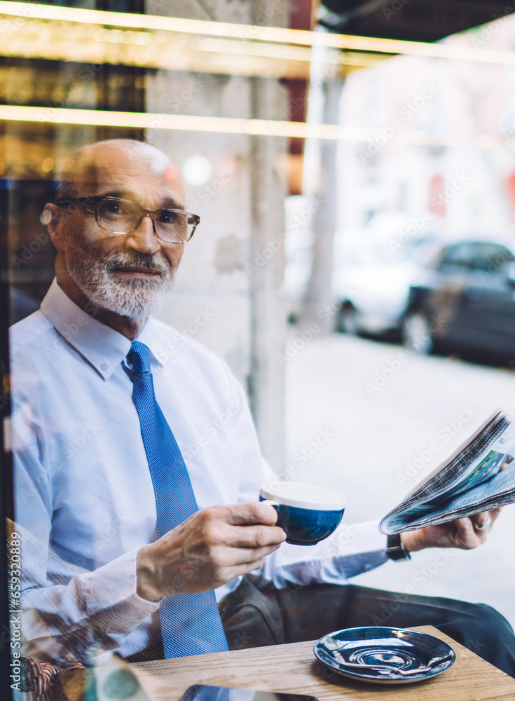Senior man in white shirt and glasses with coffee cup and newspaper looking away Senior man in white shirt and glasses with coffee cup and newspaper looking away