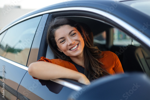 Young woman sitting in a car hand out of window. Happy woman driving a car and smiling. Portrait of happy female driver steering car with safety belt. Cute young lady happy driving car.