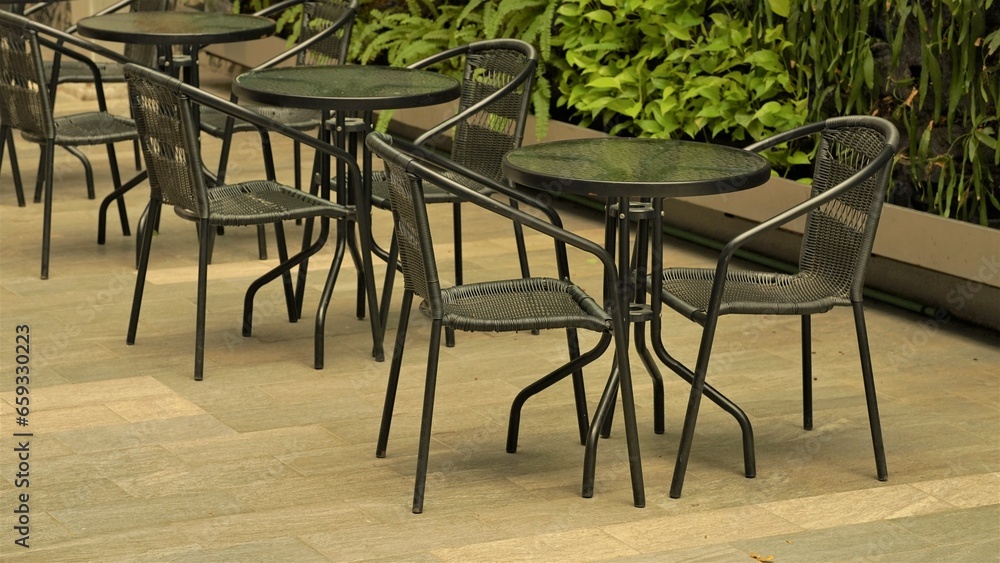 An inviting outdoor cafe setting with dark wicker chairs and matching tables, placed on a stone-tiled floor. The backdrop features lush greenery, creating a serene atmosphere ideal for relaxation