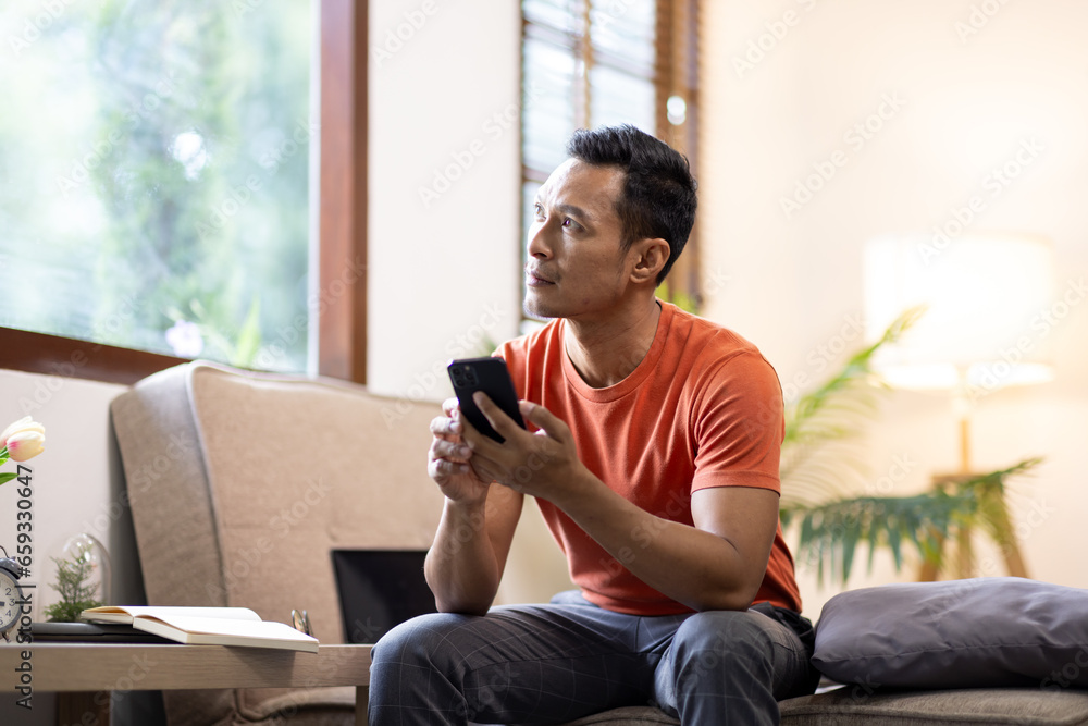 Fototapeta premium Portrait of an attractive smiling young bearded asian man wearing casual clothes sitting on a couch at the living room, using mobile phone