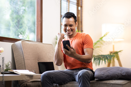 Portrait of an attractive smiling young bearded asian man wearing casual clothes sitting on a couch at the living room, using mobile phone