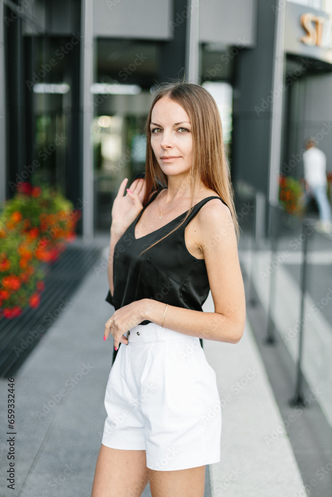 A girl with long blond hair, wearing a black top, black jacket and white shorts, poses against the backdrop of modern architecture. Young woman walking in a park with landscape design A girl with long blond hair, wearing a black top, black jacket and white shorts, poses against the backdrop of modern architecture. Young woman walking in a park with landscape design