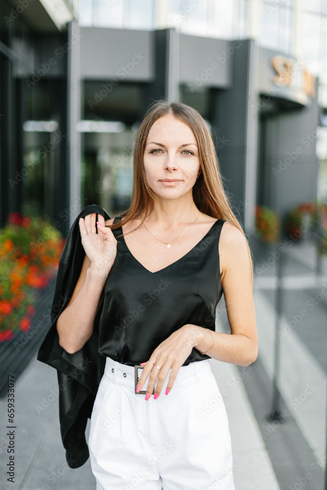 Fototapeta premium A girl with long blond hair, wearing a black top, black jacket and white shorts, poses against the backdrop of modern architecture. Young woman walking in a park with landscape design