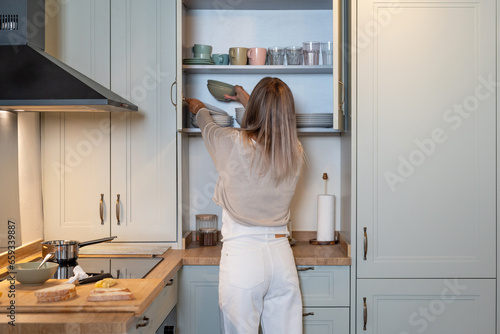 Back view of Caucasian woman in casual outfit standing at cupboard in modern kitchen