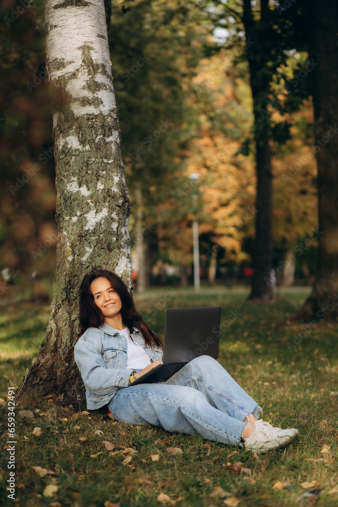 Woman sitting under the tree in park working on laptop and drinking coffee Woman sitting under the tree in park working on laptop and drinking coffee