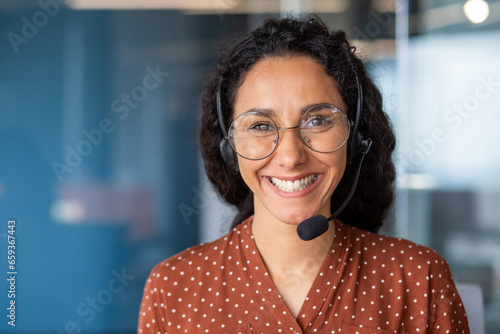 Close-up portrait of a woman with a headset, an online customer support worker smiling and looking at the camera, a business woman advising clients, working inside the office, using video call
