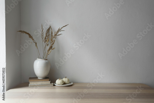 Elegant moody Scandinavian interior. Vase with dry grass bouquet, old books and little white pumpkins. Empty wall background. Modern living room, home office. Minimal autumn decor in cozy apartment.