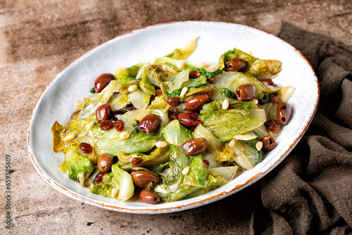 Dish with Italian salad made with sauteed broad-leaved endive or escarole with taggiasca olives, pine nuts, raisins. Vegan dish. Brown table surface. photo