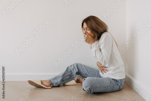 Asian Thai woman feel sick and queasy, cover mouth, has a stomachache cramp, grabbing stomach with one hand and  in white sweater, sitting on floor in white apartment room in winter.