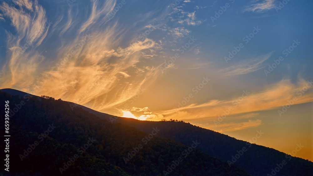 A dark forested mountainside against a dramatic evening sky with clouds glowing in the sunset light
