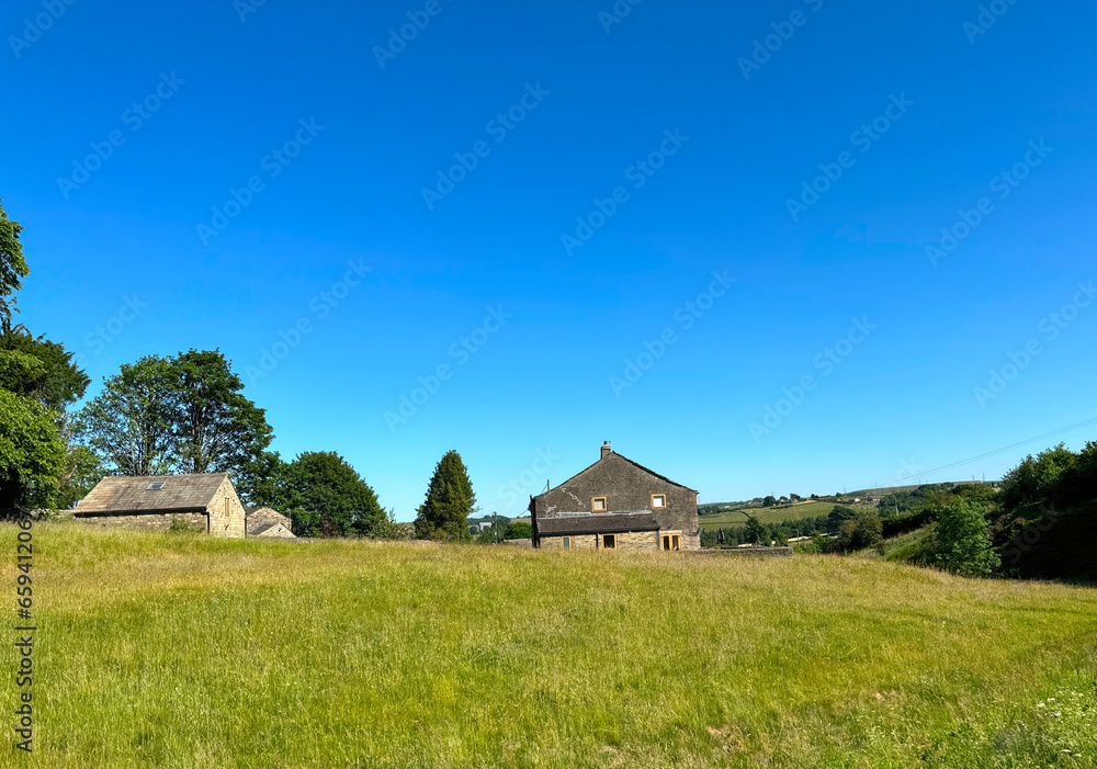 Landscape, with long grass, stone buildings, trees and hills, set against a vivid blue sky in, Ripponden, UK