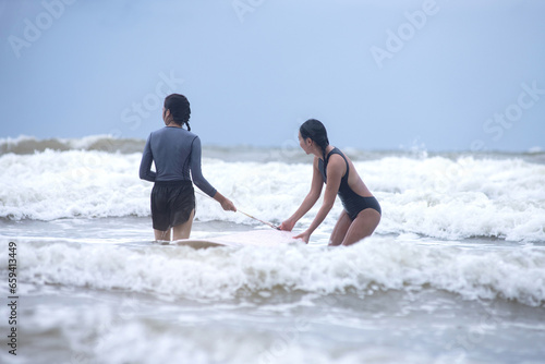 Pretty teen girl and her instructor with a surfboard go into the water waiting for waves suitable for surfing, concept of water sports activities