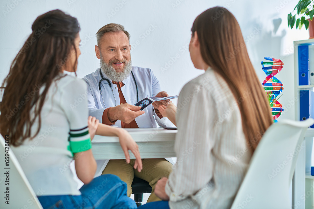 Obraz premium cheerful grey bearded doctor showing lgbt couple their ultrasound, in vitro fertilization concept