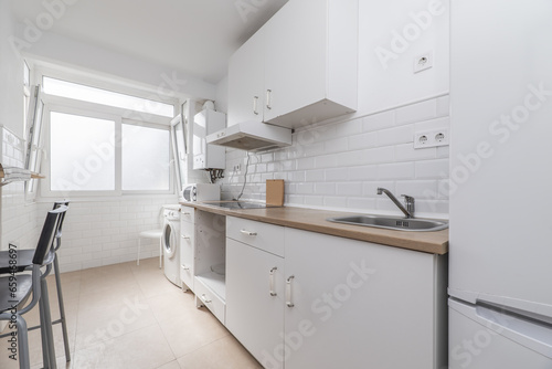 Furnished kitchen on one of the walls with white furniture, integrated white appliances, high stools on one side and a light wood-colored countertop and a white aluminum window