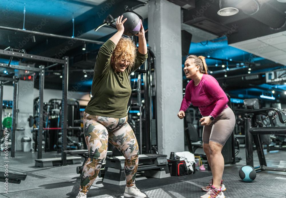 Two plus-size women workout and exercise with medicine ball at the gym. They're determined to achieve their goals and inspire others along the way.	 - obrazy, fototapety, plakaty 