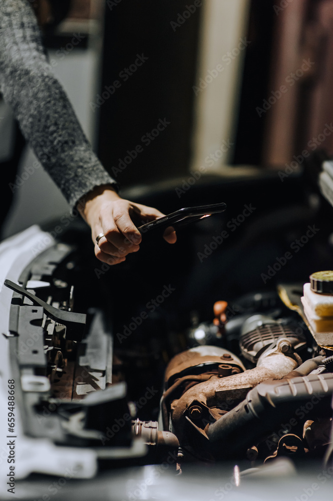 A woman mechanic repairs a car with a breakdown in the engine under the hood using tools at night, highlighting it with a flashlight on her phone or smartphone. Photography, portrait.