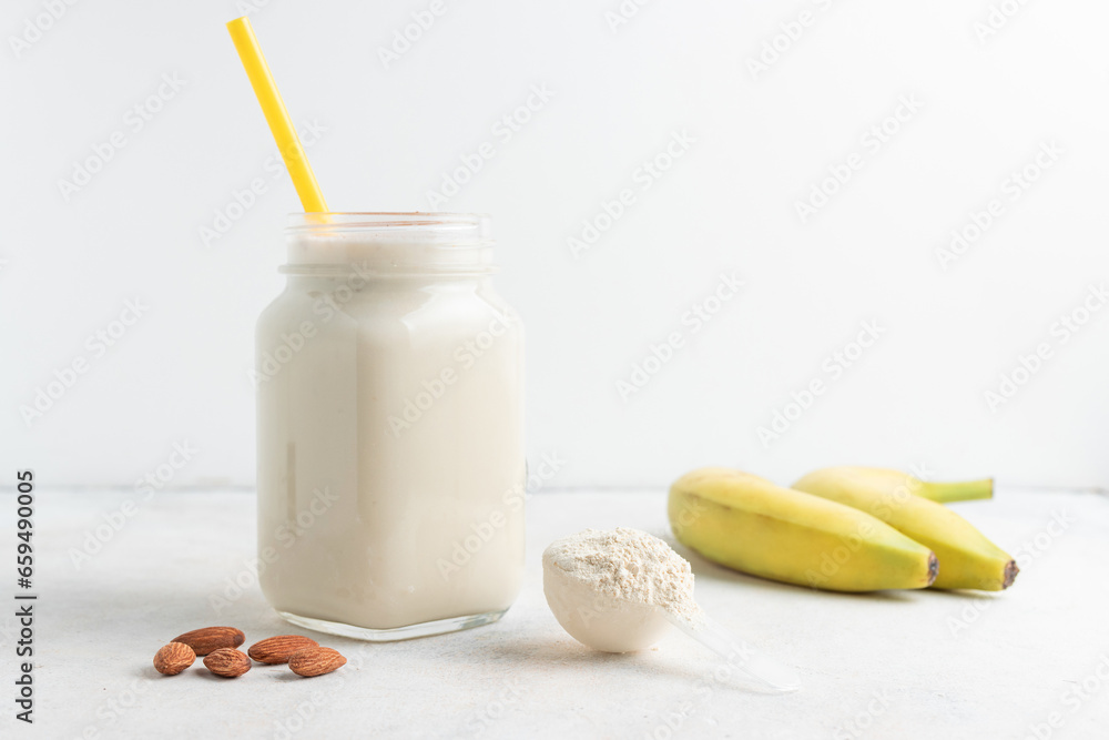 Glass jar of protein milkshake drink or smoothie and whey protein powder in measuring spoon, bananas, almond nuts on a white background. sport nutrition, bodybuilding food supplements Glass jar of protein milkshake drink or smoothie and whey protein powder in measuring spoon, bananas, almond nuts on a white background. sport nutrition, bodybuilding food supplements