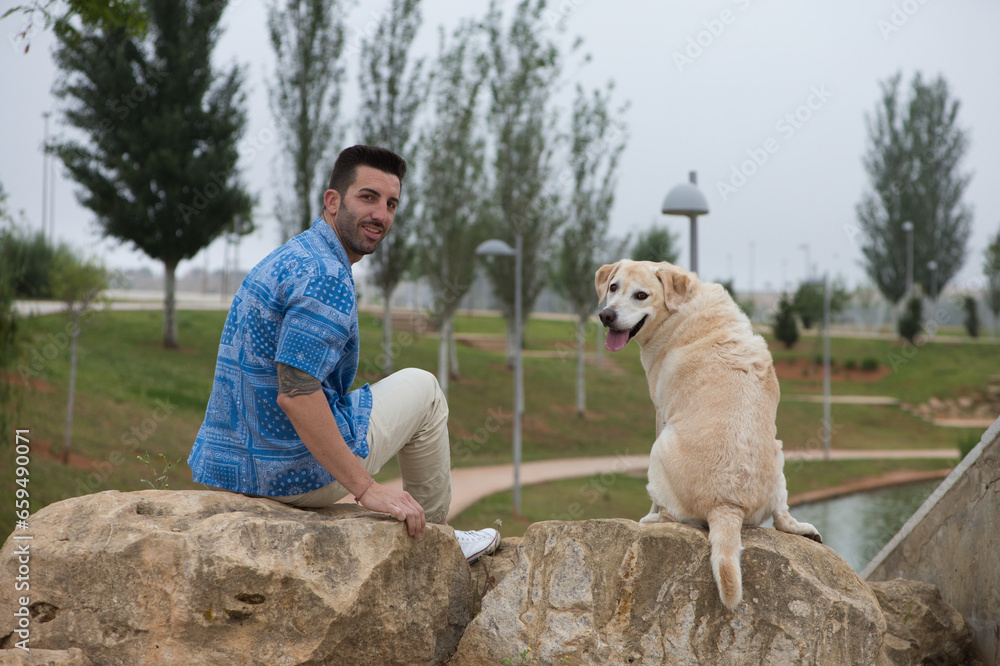 Handsome young man with beard and his large dog sitting on rocks looking back. The dog is a brown Labrador retrieve. Concept pets and companion animals. International dog day Handsome young man with beard and his large dog sitting on rocks looking back. The dog is a brown Labrador retrieve. Concept pets and companion animals. International dog day