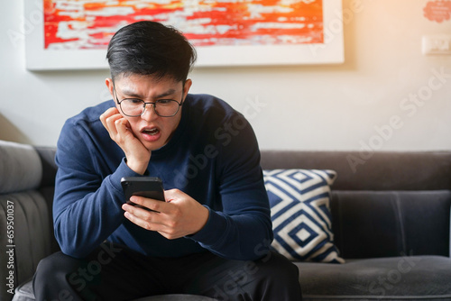 close up young asian man frowned on face with shock feeling while using smartphone to reading about fake news and rumor in home for people lifestyle concept photo
