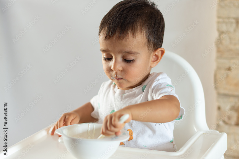 Toddler with spoon in hand and dirty mouth learns to eat while looking at his plate Toddler with spoon in hand and dirty mouth learns to eat while looking at his plate