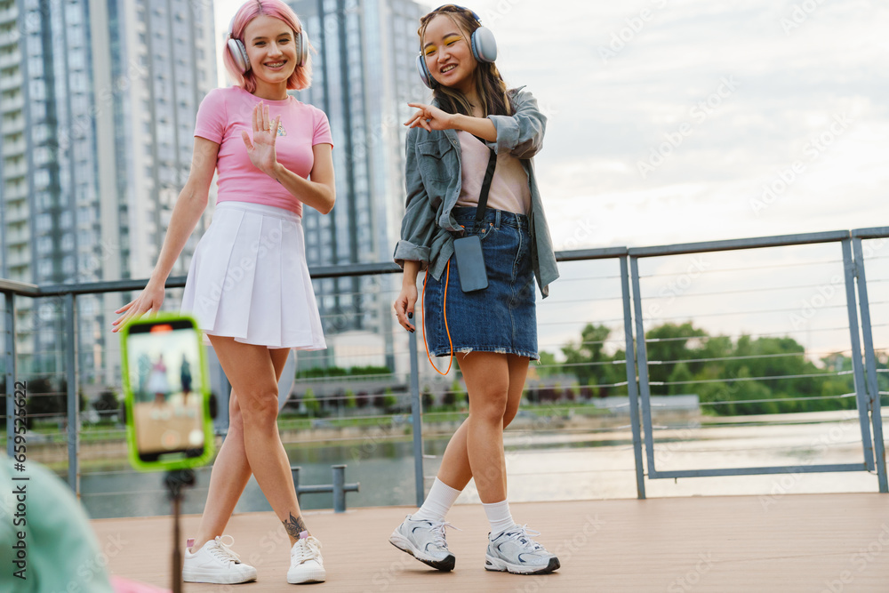 Obraz premium Two cheerful girls making video with smartphone while standing on city waterfront