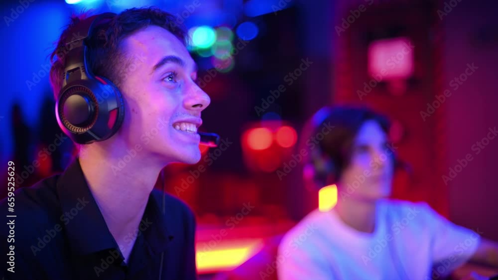 Two teen friends in headphones are talking and playing a game console using gamepads, talking and laughing while sitting on bean bags. Blue and red illumination Two teen friends in headphones are talking and playing a game console using gamepads, talking and laughing while sitting on bean bags. Blue and red illumination
