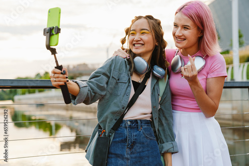 Two girls taking self portraits with smartphone and selfie stick while standing on city waterfront